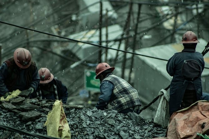 Several miners working outdoors in a quarry with buildings and electrical lines in the background.