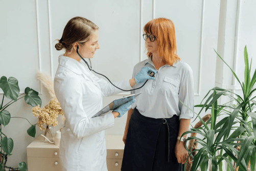Healthcare professional using a stethoscope to examine a patient in a medical office and documenting it digitally on a tablet.