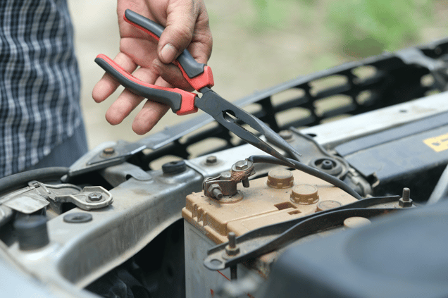 Close-up of hands using red-handled pliers to work on a car battery terminal in an engine compartment.