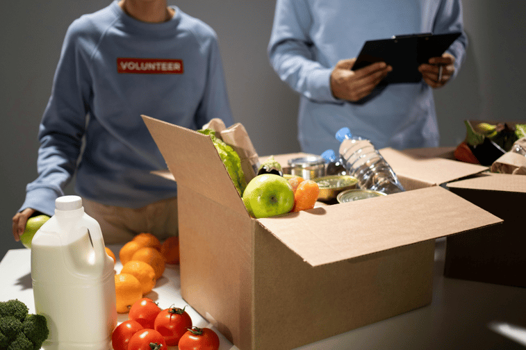 Two volunteers in blue sweatshirts are reviewing food in cardboard boxes and taking notes.