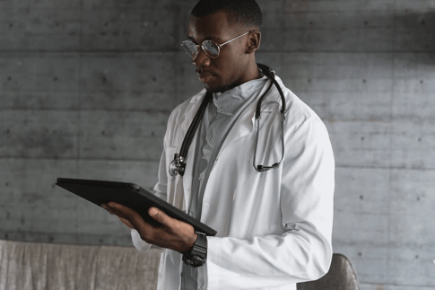 A doctor in a white coat, holding a tablet, stands in a professional, modern clinical setting.