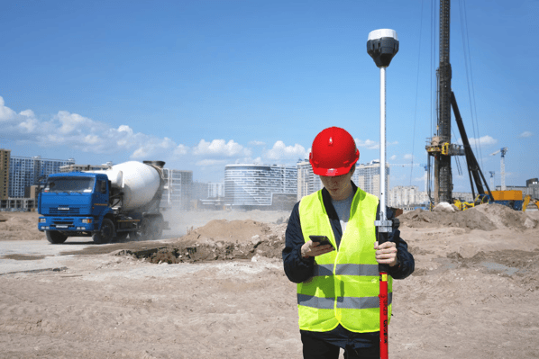A construction inspector in a red hard hat and yellow safety jacket enters inspection data into a mobile app on his smartphone