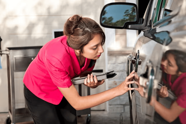 Woman inspecting scratches on a car door while holding a clipboard and taking notes.