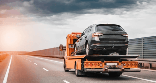 Black SUV loaded onto yellow tow truck driving down an empty highway under a cloudy sky, symbolizing tow truck dispatch and roadside assistance services supported by mobile apps.