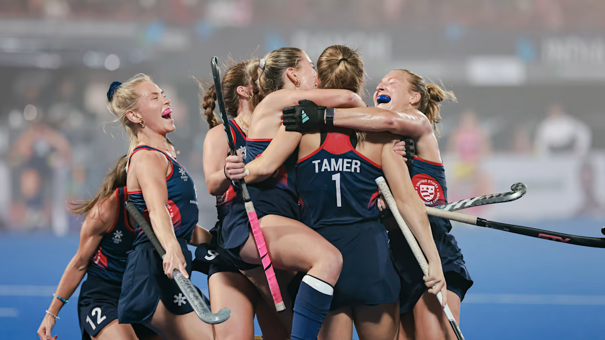 Women’s field hockey team in navy blue uniforms, celebrating with hugs and cheers on the field after a victory