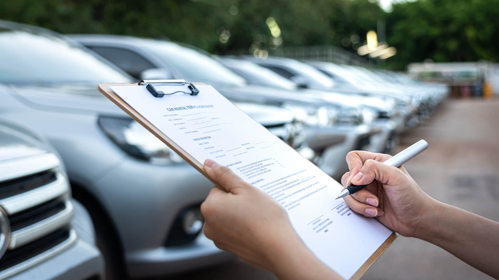 Person holding a clipboard and filling out a car rental or inspection form in front of a row of parked silver cars